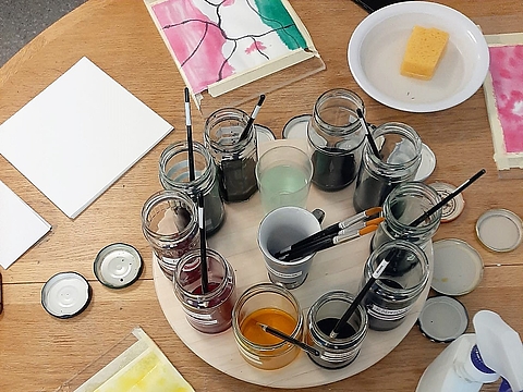 Auf einem Tisch stehen verschiedene flüssige Naturfarben in Schraubgläsern in der Mitte. Rings herum liegen Pinsel und angefangene Bilder, die mit diesen Farbe gemalt werden.