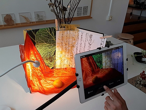 Auf einem Tisch wurde die Innenseite eines Pappkartons mit bedruckten Papieren und verschiedenen Stoffen zu einer Szenerie gestaltet. Eine Klemmlampe leuchtet das Arrangement aus. Im Vordergrund ist ein Tablet, mit dem gerade ein Foto von diesem Arragement gemacht wird.