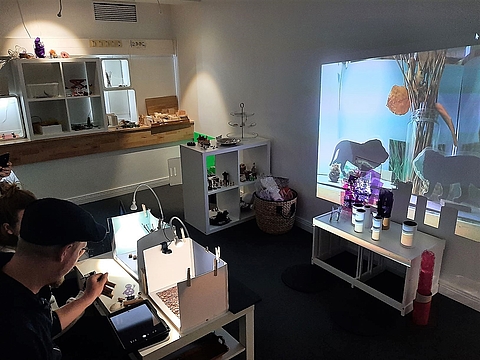 Zwei erwachsene Personen sitzen an einem Tisch und haben in Pappkartons verschiedene Szenerien gestaltet. Diese werden mit Tablets aufgenommen und mit einem Beamer groß an die gegenüberliegende Wand projeziert. Zu erkennen sind verschiedene Muster und Silhouetten von Spielzeugtieren. 
